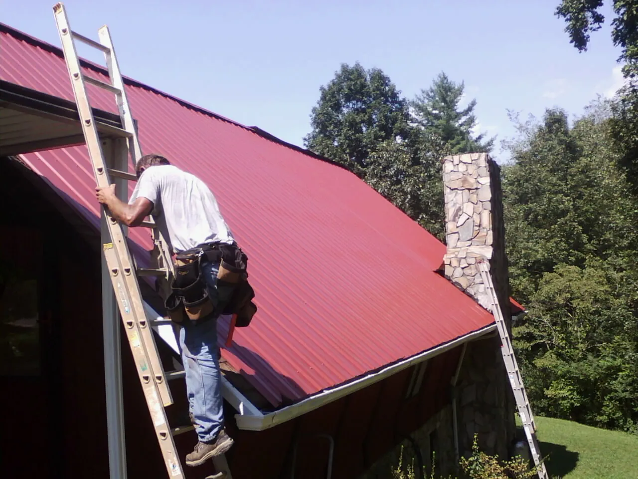 Roofer on a ladder beside a red metal roof and stone chimney — Soffit & Fascia Repair by Roofing Brothers in Smithville Flats